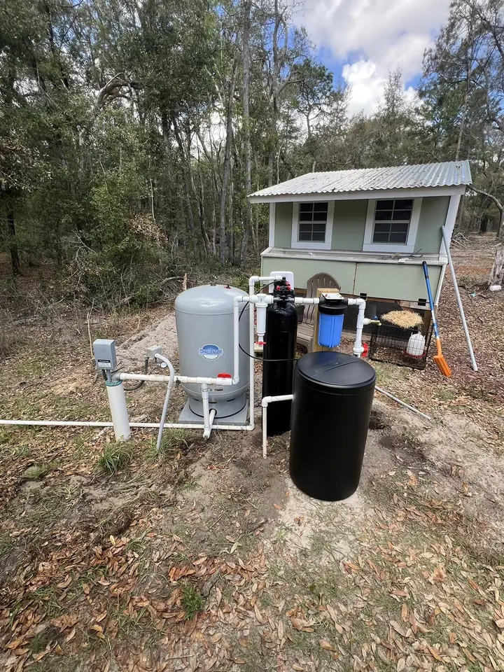 A new gray pressure tank, black softener tank, blue filter housing and brine tank plumbed in PVC beside a shed under live oaks